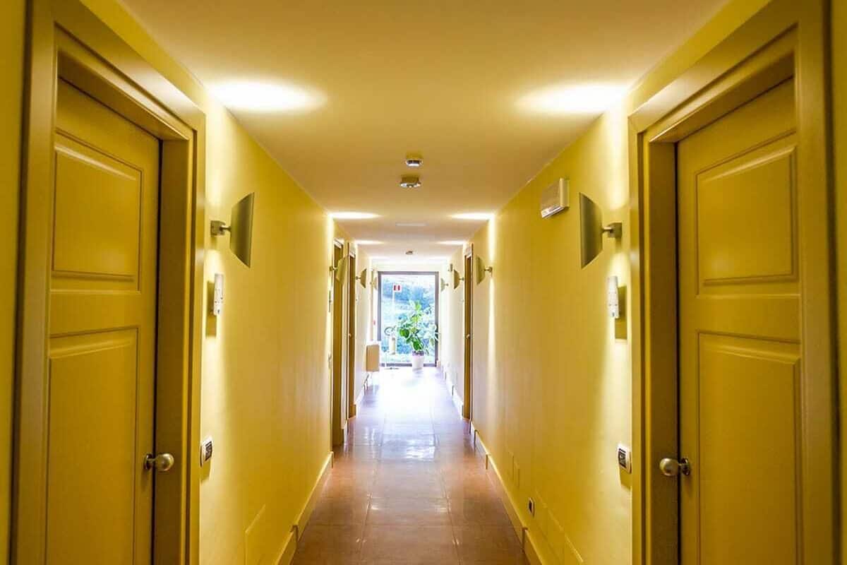 A long, narrow hallway with yellow walls and doors, leading to a bright room with a window.