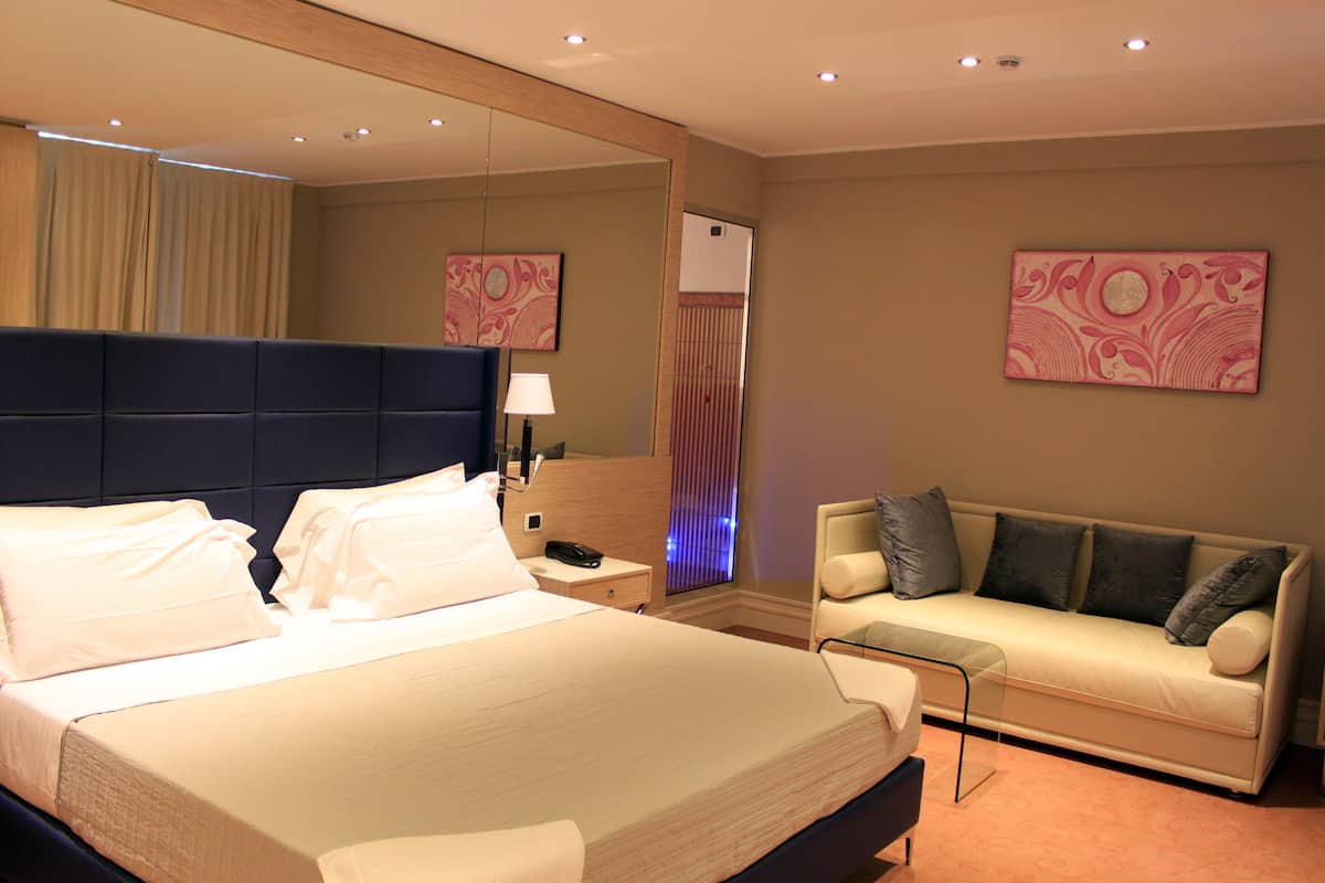 A hotel room with a large bed, a sofa, a nightstand, a lamp, and two wall-mounted artworks.