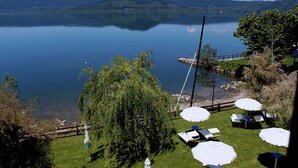 A lakeside view with a dining area featuring tables and umbrellas, surrounded by greenery and a wooden deck. - Relais Sans Souci and Spa (Ronciglione)