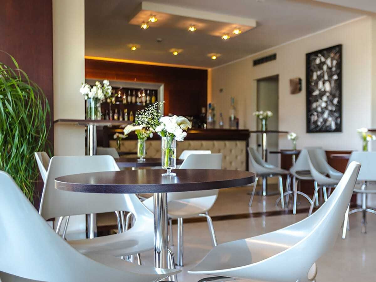 A modern hotel lobby with round tables and white chairs, a bar area in the background, and a large abstract painting on the wall.