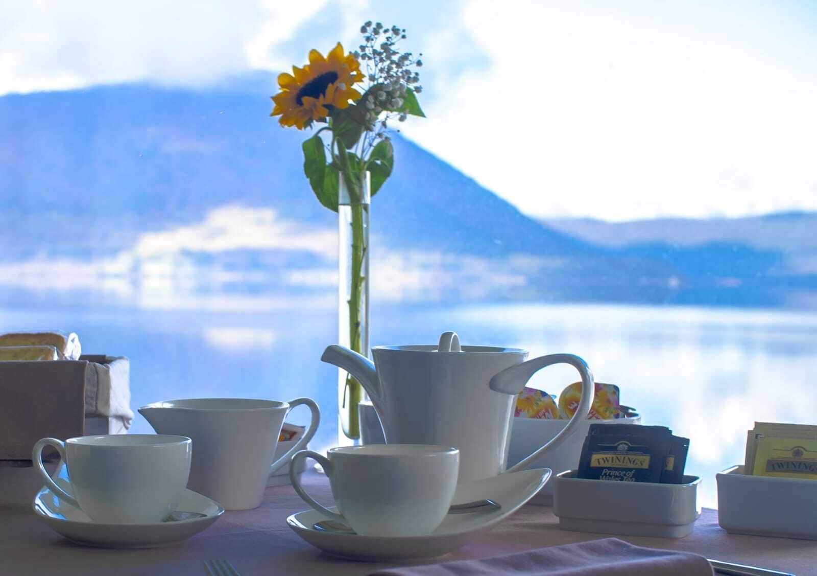 Una mesa con un juego de té, un florero con un girasol y vistas a un lago y montañas.