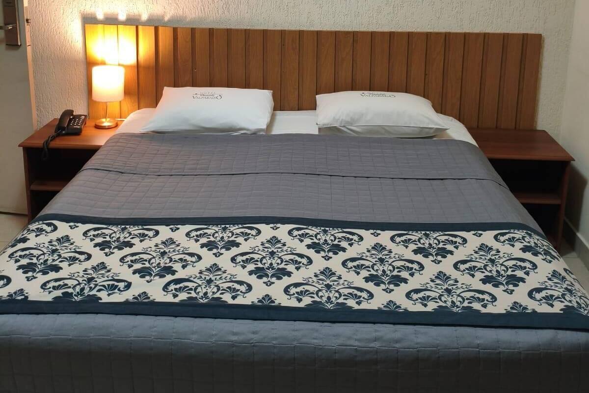 A hotel room with a wooden headboard, a bed with patterned bedding, a bedside lamp, and a wall-mounted artwork of small vases.