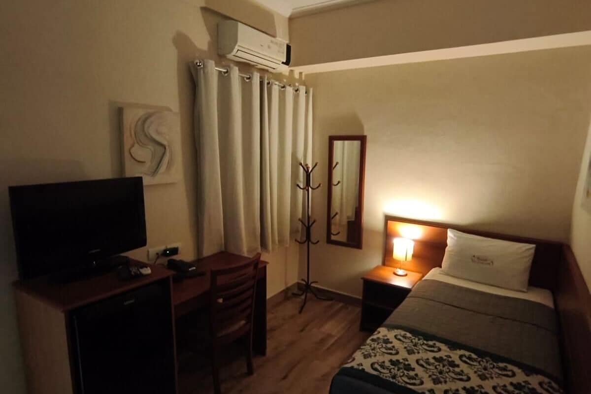 Quarto de hotel com cama, escrivaninha, televisão, cabideiro e abajur.