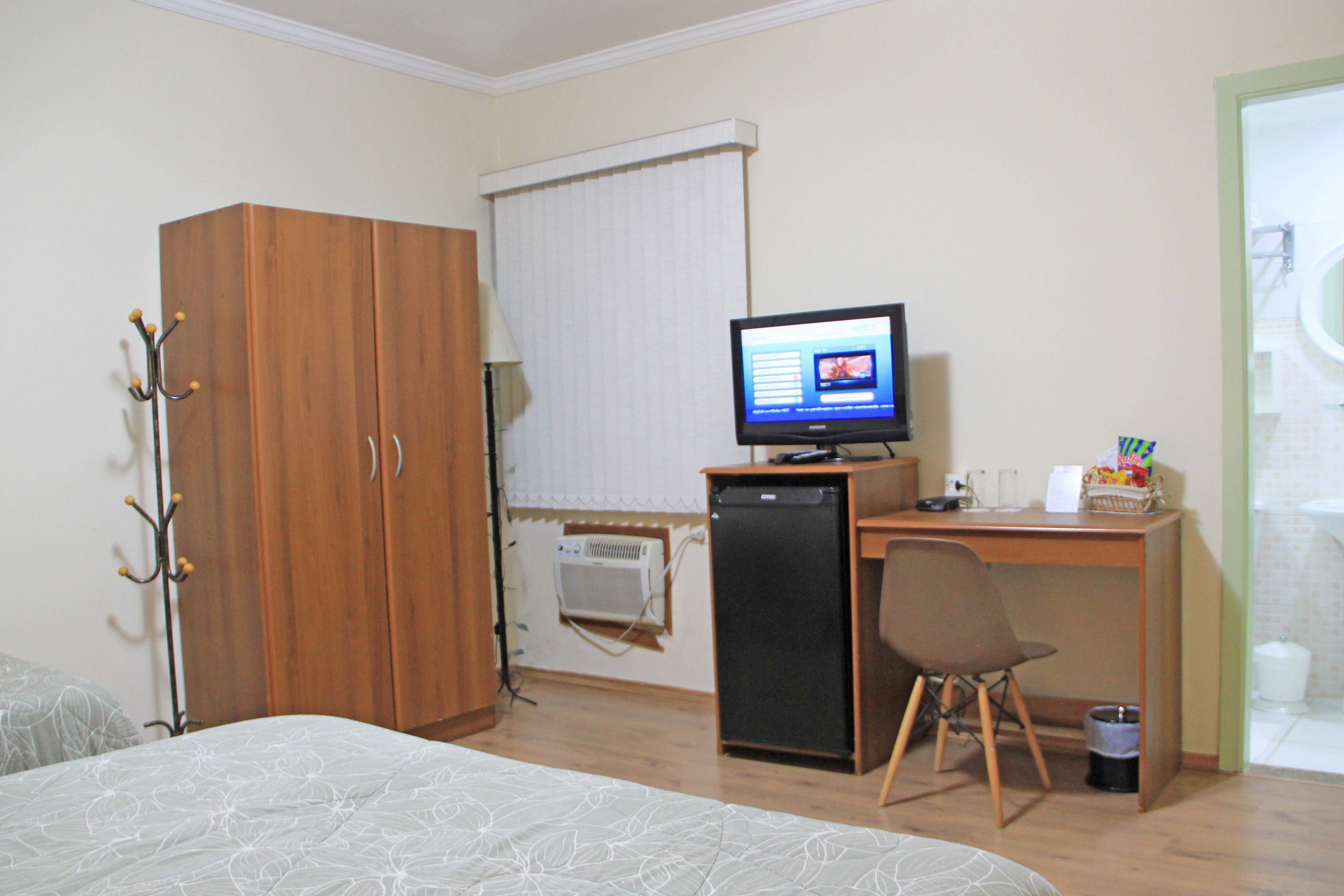 Ein Schlafzimmer mit Bett, Schreibtisch mit Computer, Stuhl, Kleiderschrank und einem Fenster mit Jalousien.