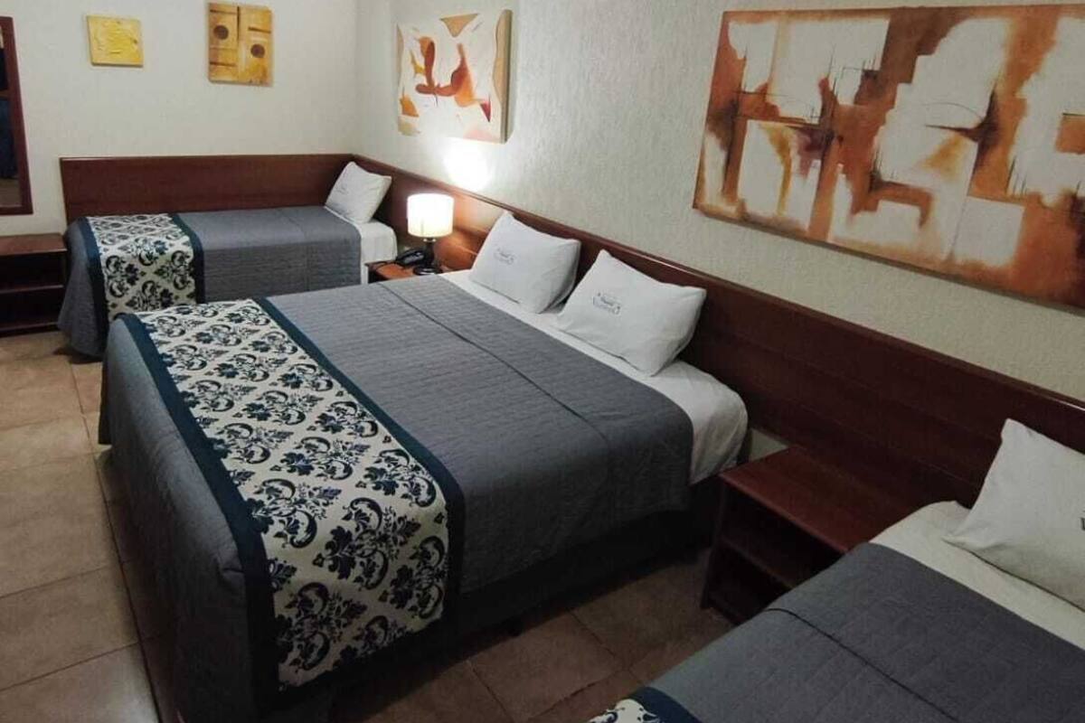 A hotel room with three beds, each with a patterned bedspread, a bedside table with a lamp, and artwork on the walls.