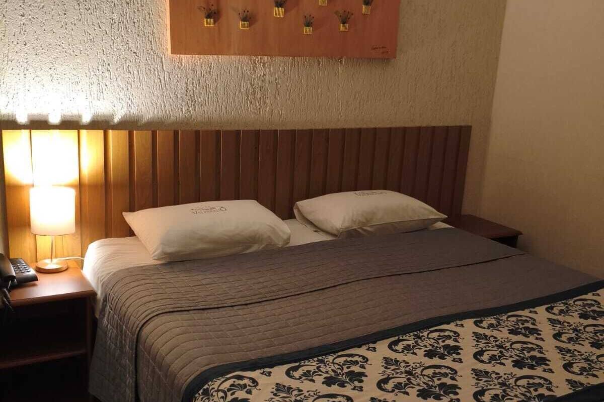 A hotel room with a wooden headboard, a bed with two pillows, a bedside table with a lamp, and a wall-mounted light fixture.
