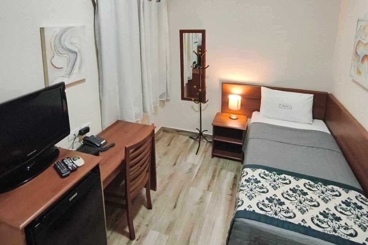A hotel room with a bed, a desk with a TV, a chair, a bedside table with a lamp, and a wardrobe.