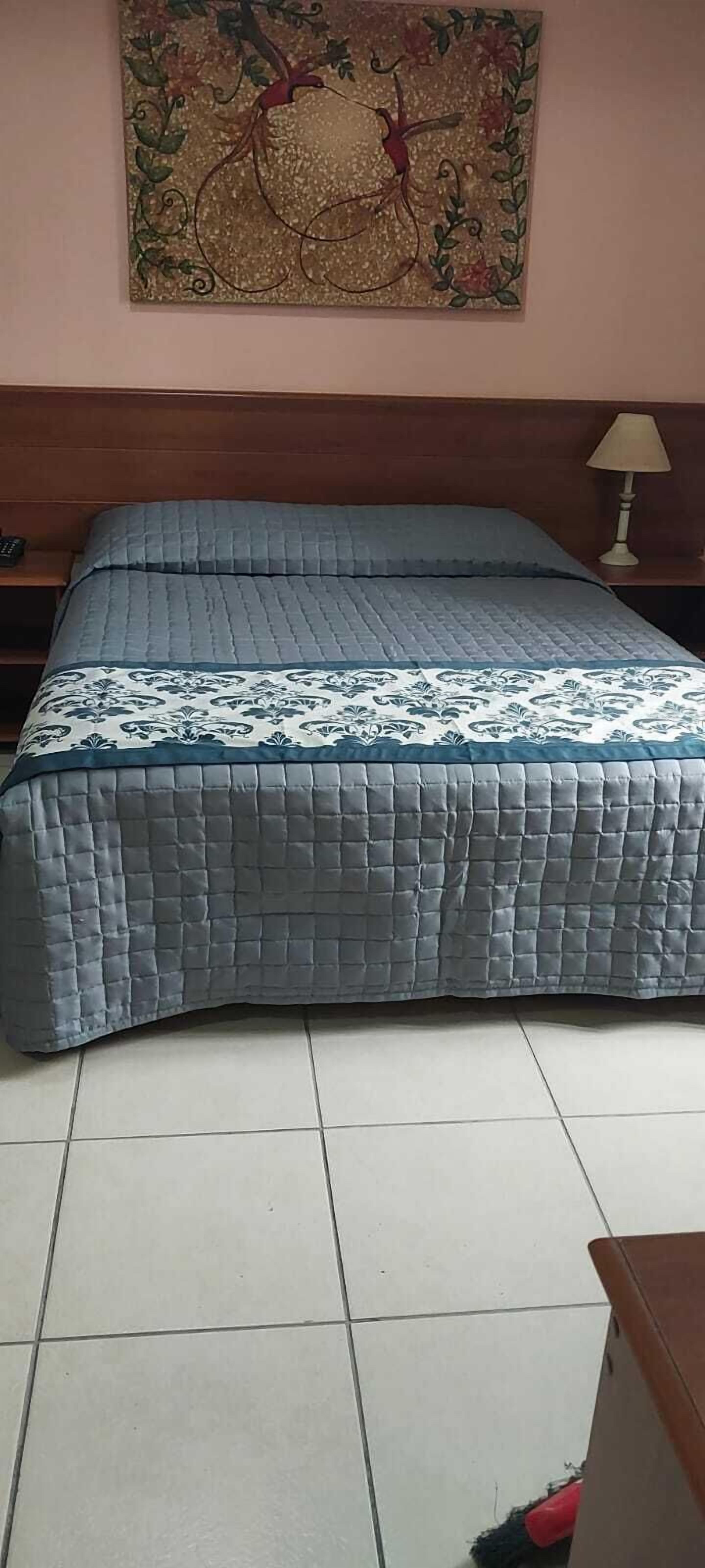 Un dormitorio con cama, mesita de noche, lámpara y un cuadro en la pared.
