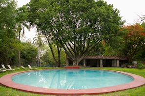 Una piscina circular rodeada de césped y árboles, con un pabellón al fondo.