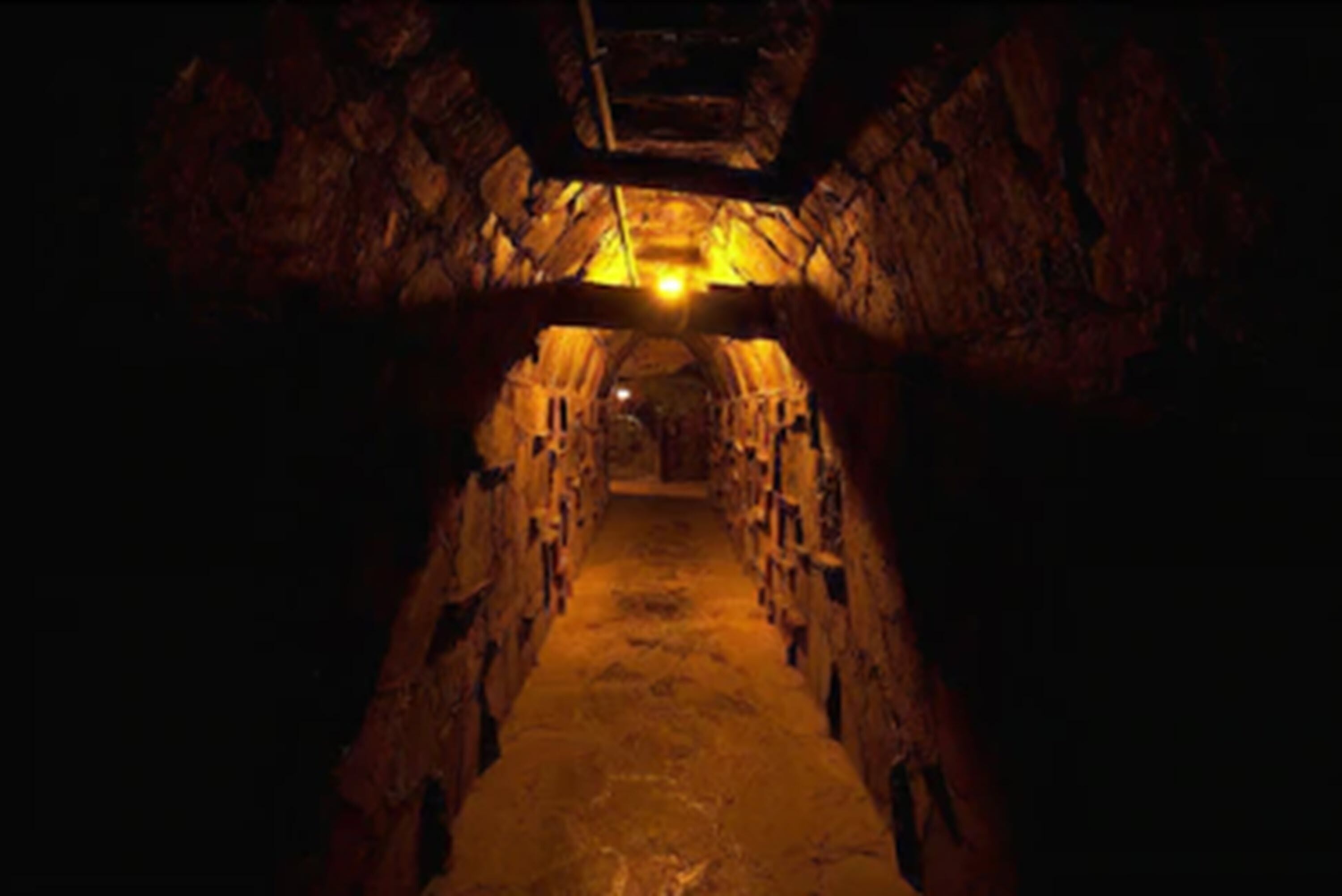 A narrow, dimly lit tunnel with stone walls and a wooden ceiling.