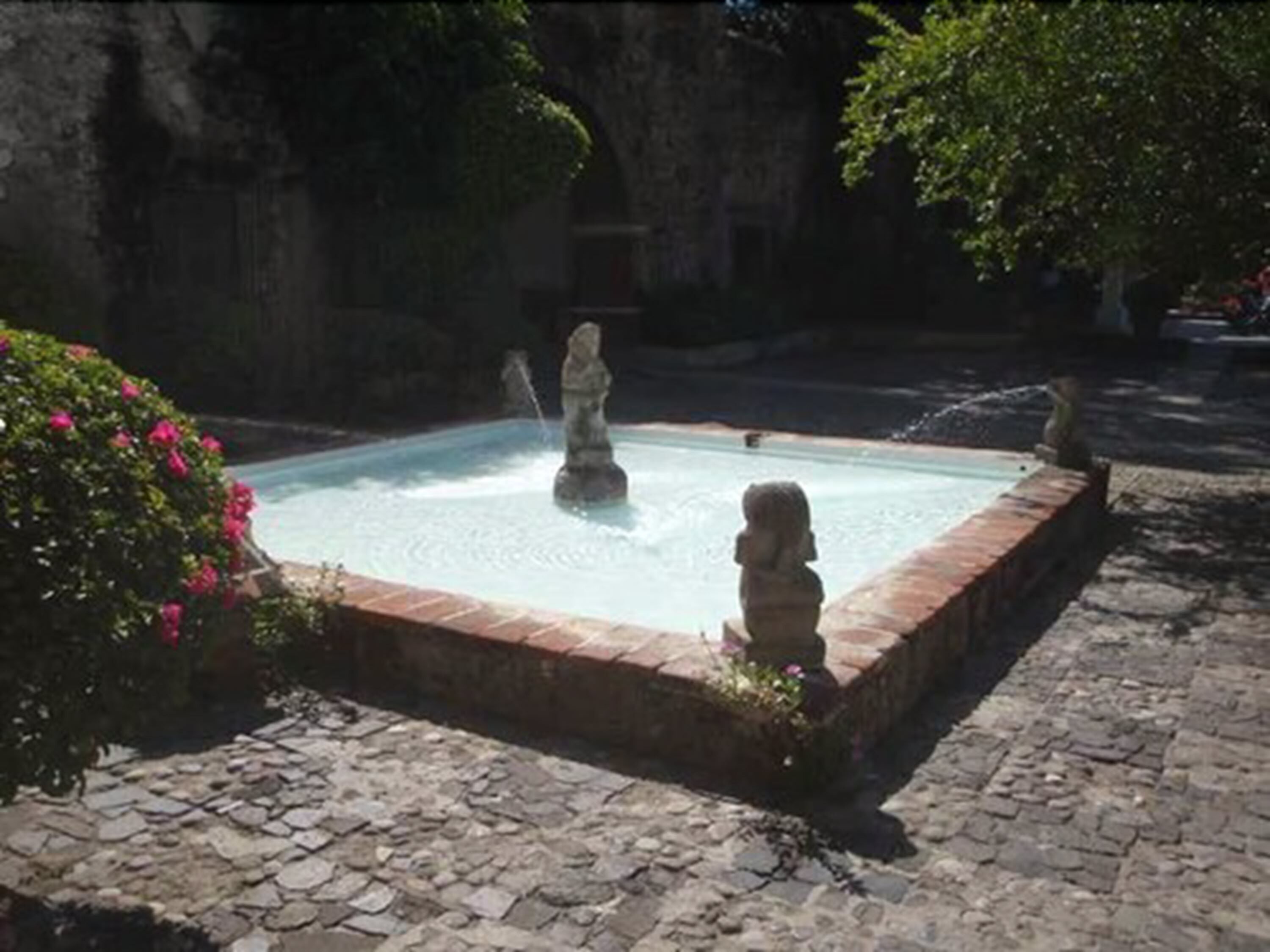 Una piscina cuadrada con estatuas de piedra, rodeada por un borde de ladrillo y suelo adoquinado.