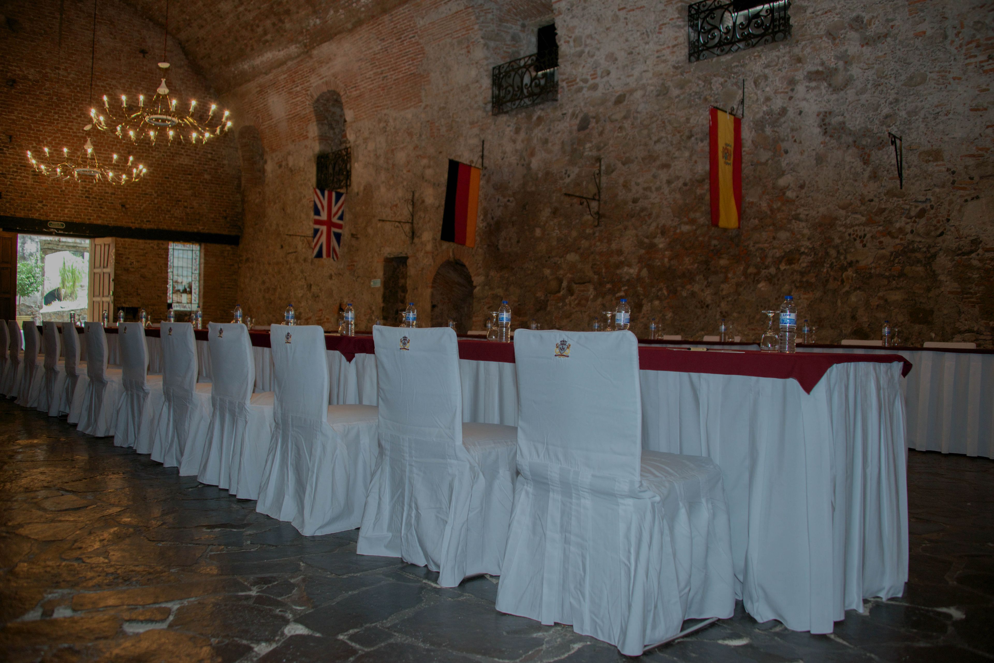 Un salón de banquetes con filas de mesas, sillas y una araña. Se observan banderas en las paredes.