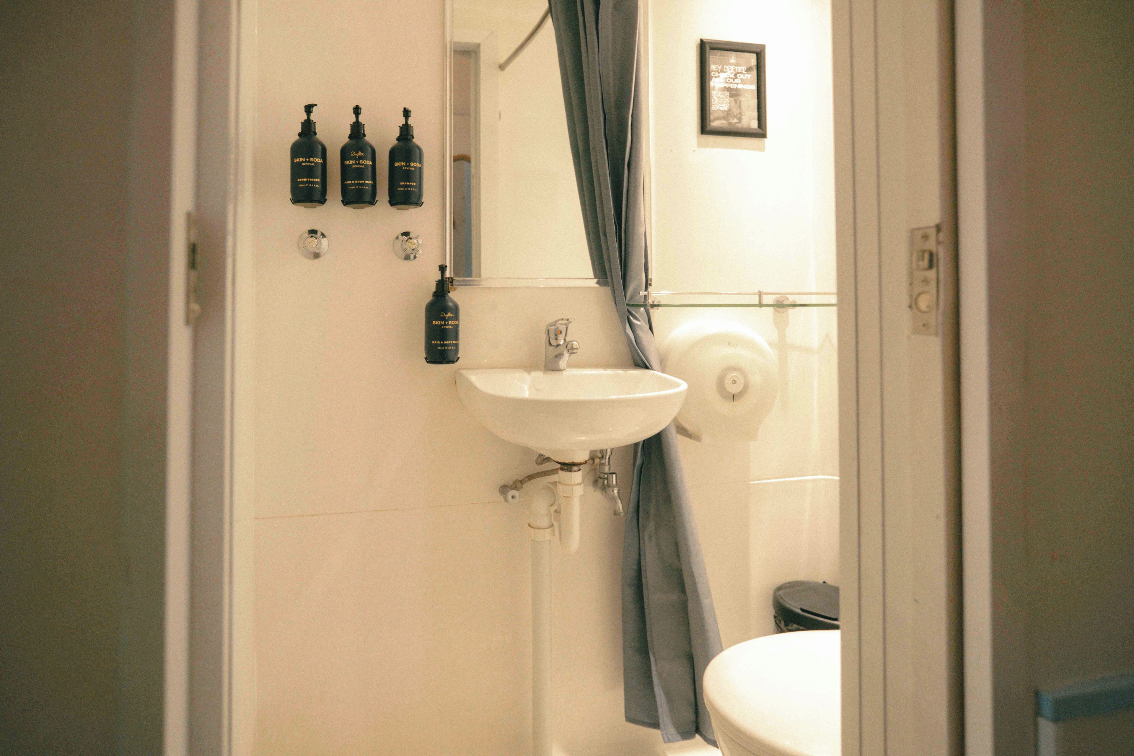 Un baño con lavamanos, cortina de ducha y tres botellas de productos para el cuidado personal y refrescos en la pared.