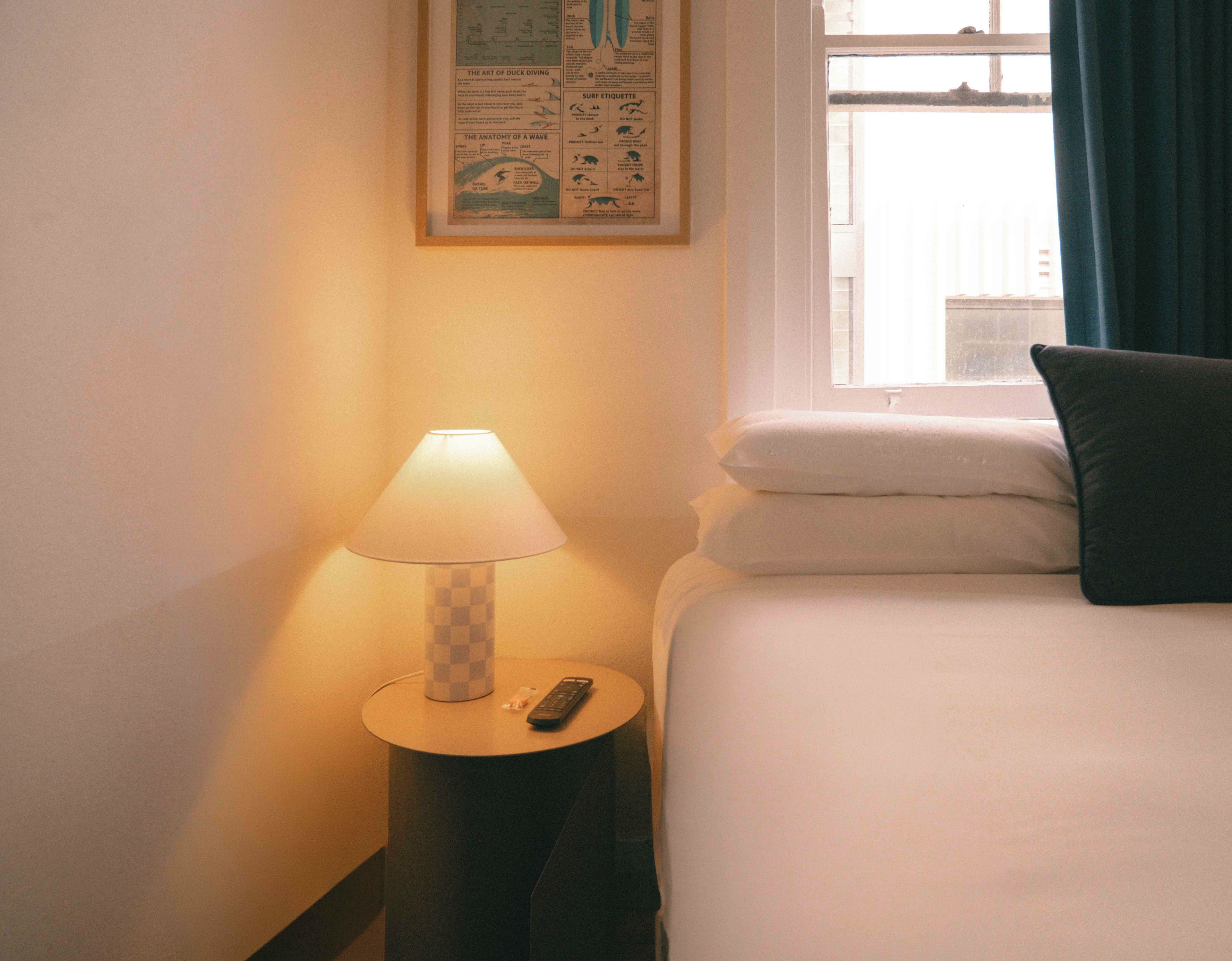 A hotel room with a bed, a nightstand with a lamp, a remote control, and a window with curtains.