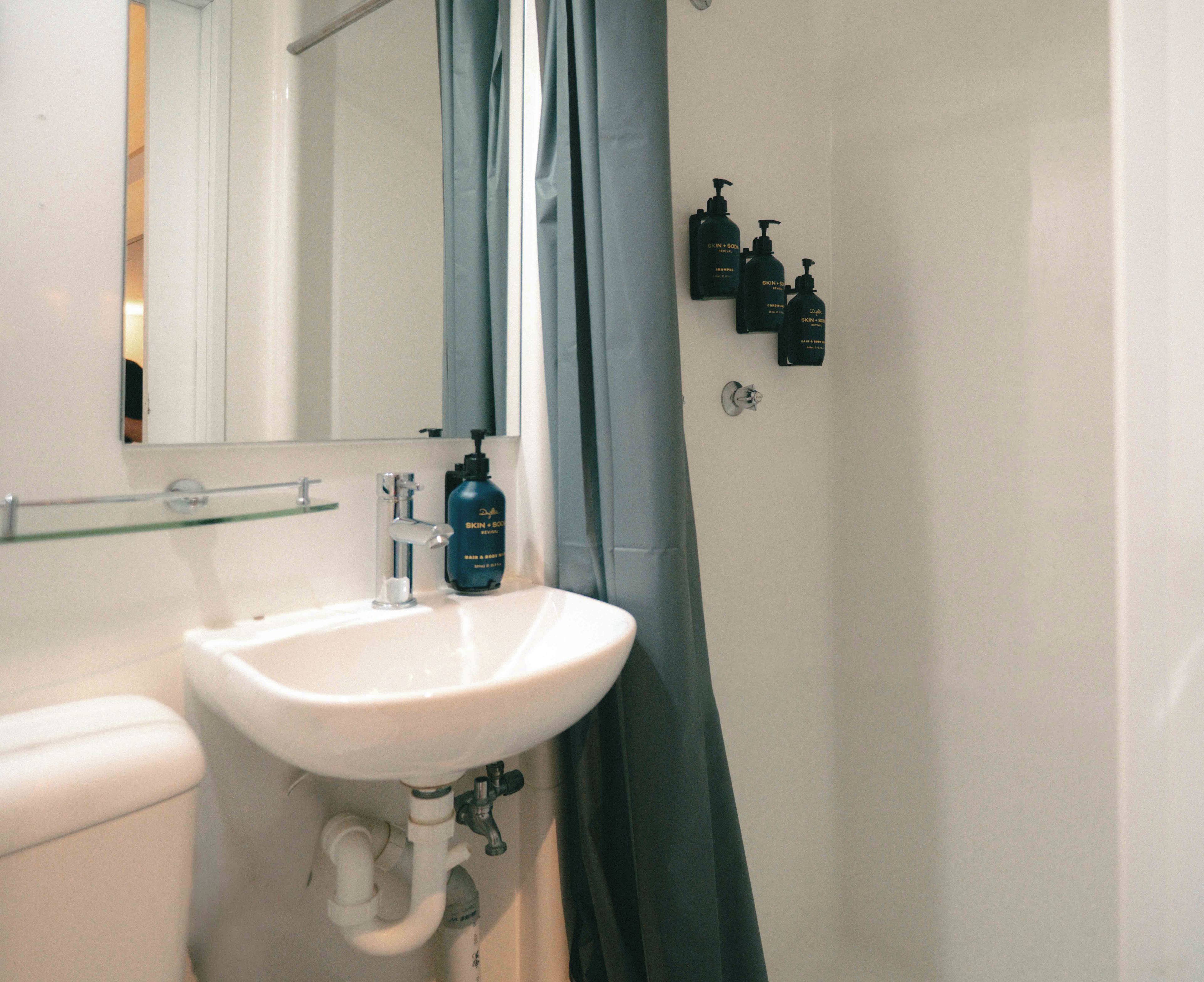 Une salle de bain avec un lavabo blanc, un miroir, un rideau de douche et plusieurs flacons de produits de soins pour la peau accrochés au mur.