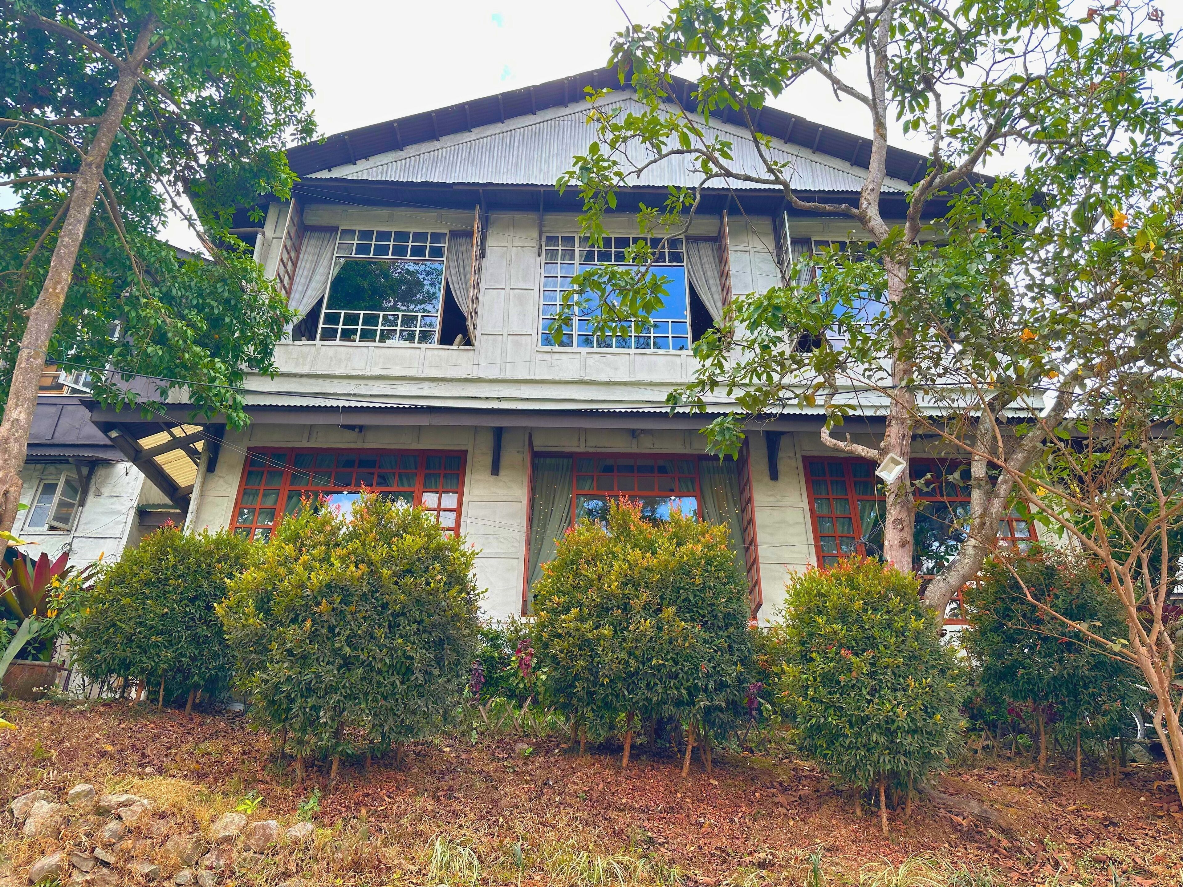 Une maison à deux étages, dotée d’un toit en tôle ondulée, de nombreuses fenêtres à encadrements rouges et d’un jardin devant la maison, agrémenté de divers arbustes et arbres.