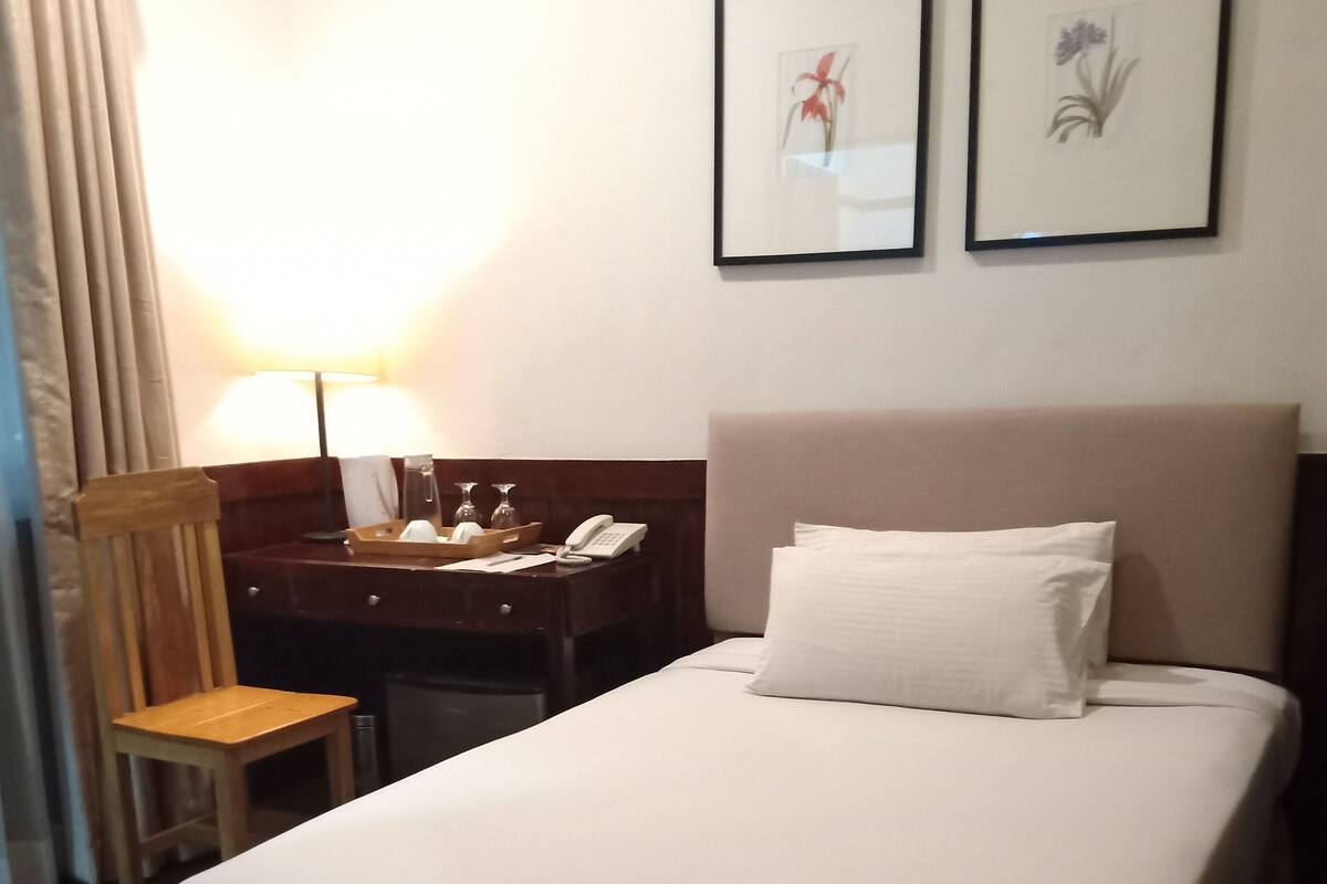A hotel room with a bed, a desk with a lamp, and two framed pictures on the wall.