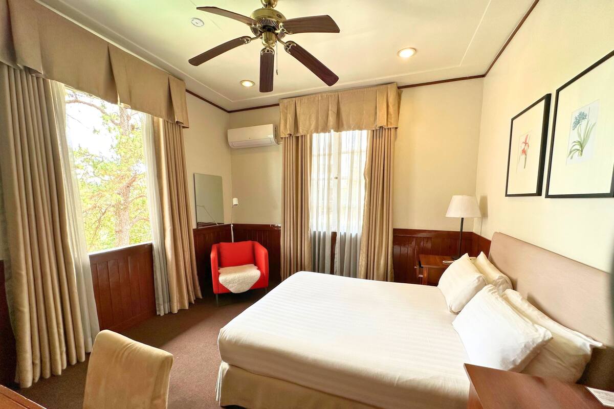 A hotel room with a large bed, a red armchair, a wooden nightstand, and a ceiling fan.