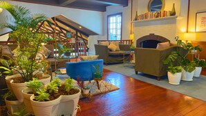 A cozy living room with a fireplace, comfortable seating, and multiple potted plants.