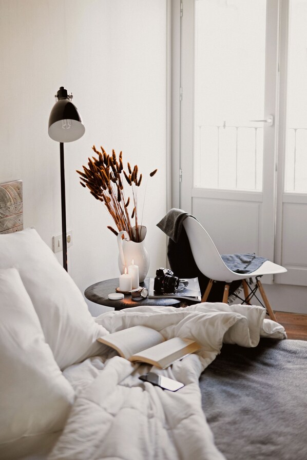 A bedroom with a bed, a bedside table with a lamp, a chair, a vase with dried plants, and a window with a view.
