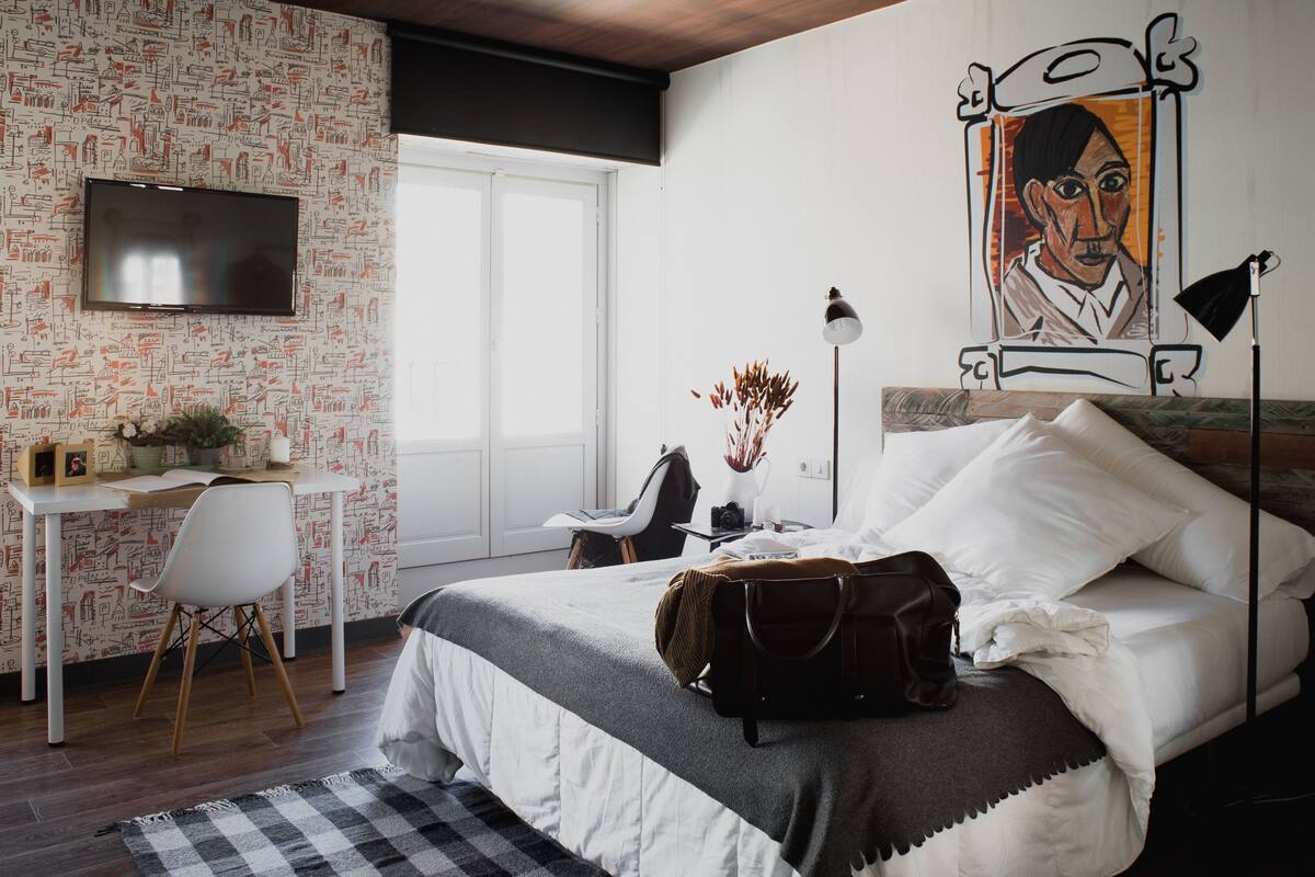 A bedroom with a bed, desk, chair, and a painting on the wall.