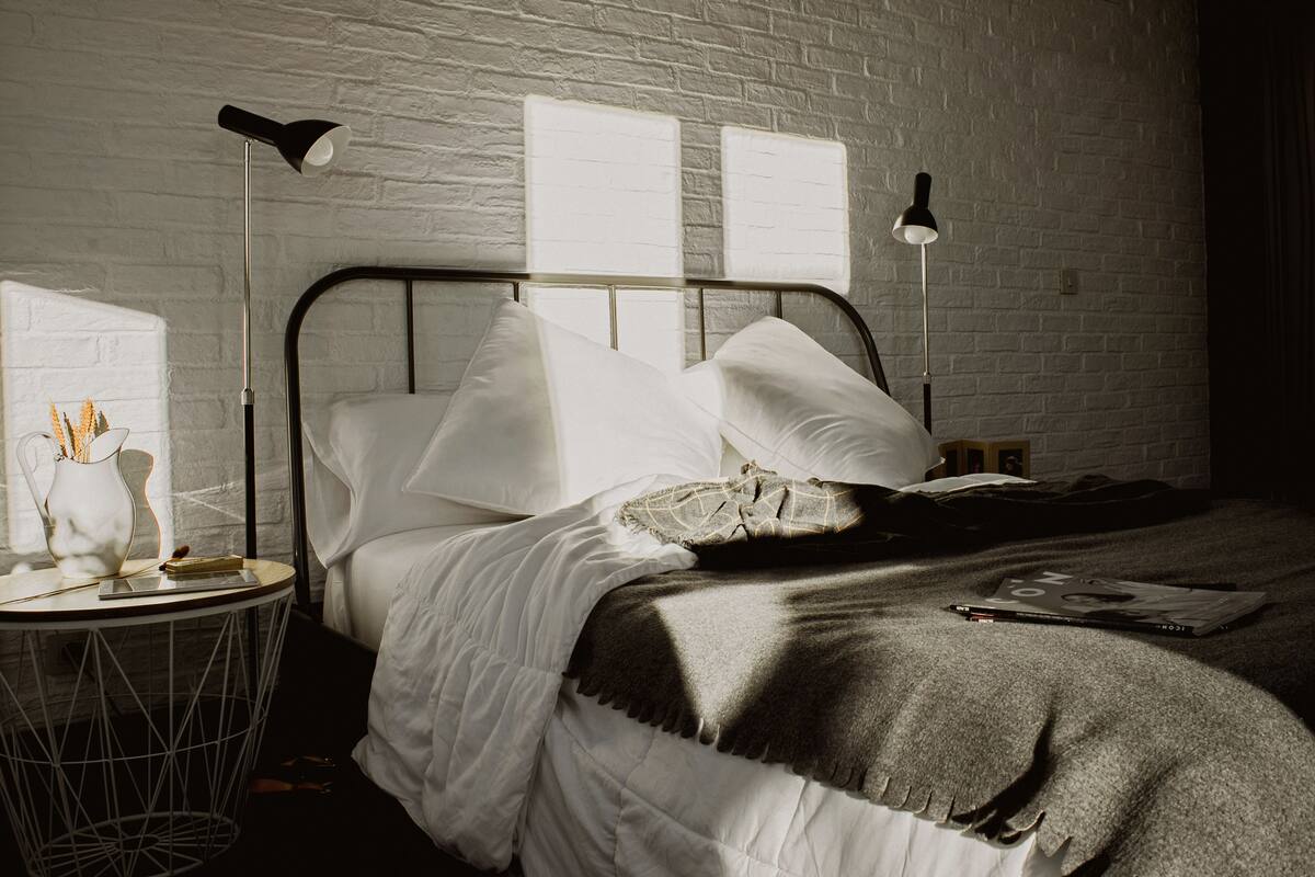 A neatly made bed with white linens and a gray throw blanket, a bedside table with a lamp, and a brick wall.