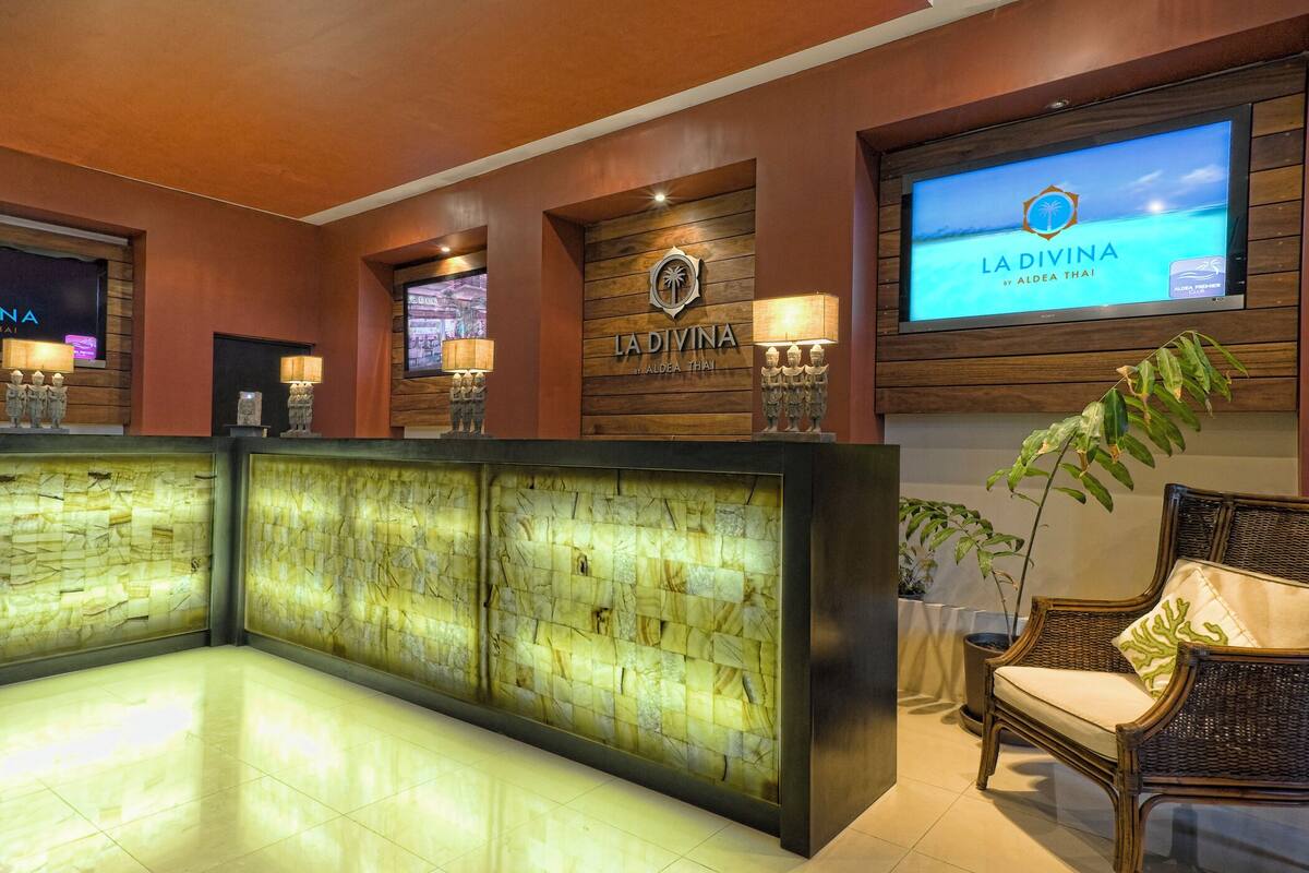 Un espace d’accueil doté d’un comptoir en bois, d’un écran de télévision diffusant « LA DIVINA by Aleda Thai » et d’un coin salon avec des meubles en osier.