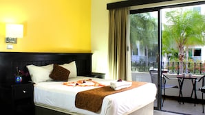 A hotel room with a bed, a nightstand, a window with curtains, a balcony with a table and chairs, and a view of greenery outside.