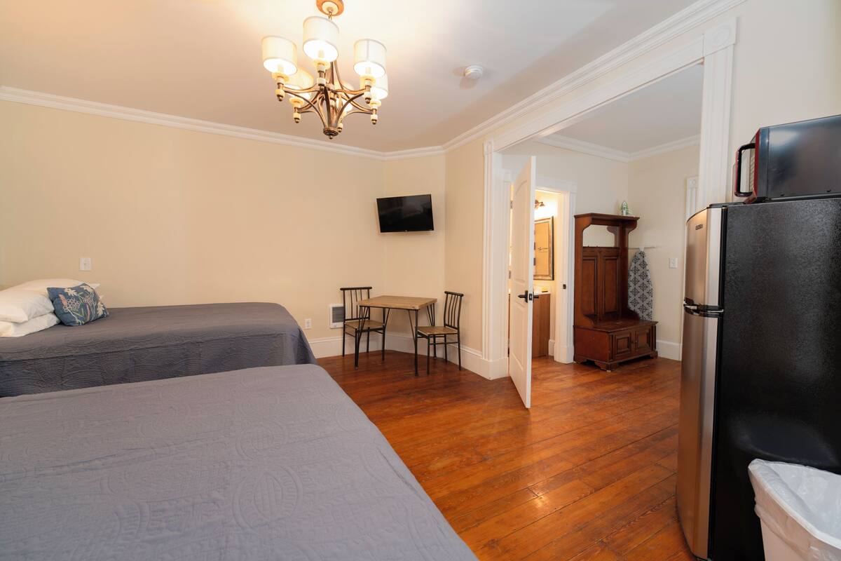 A bedroom with two beds, a dining area with a table and chairs, a TV mounted on the wall, and a refrigerator.