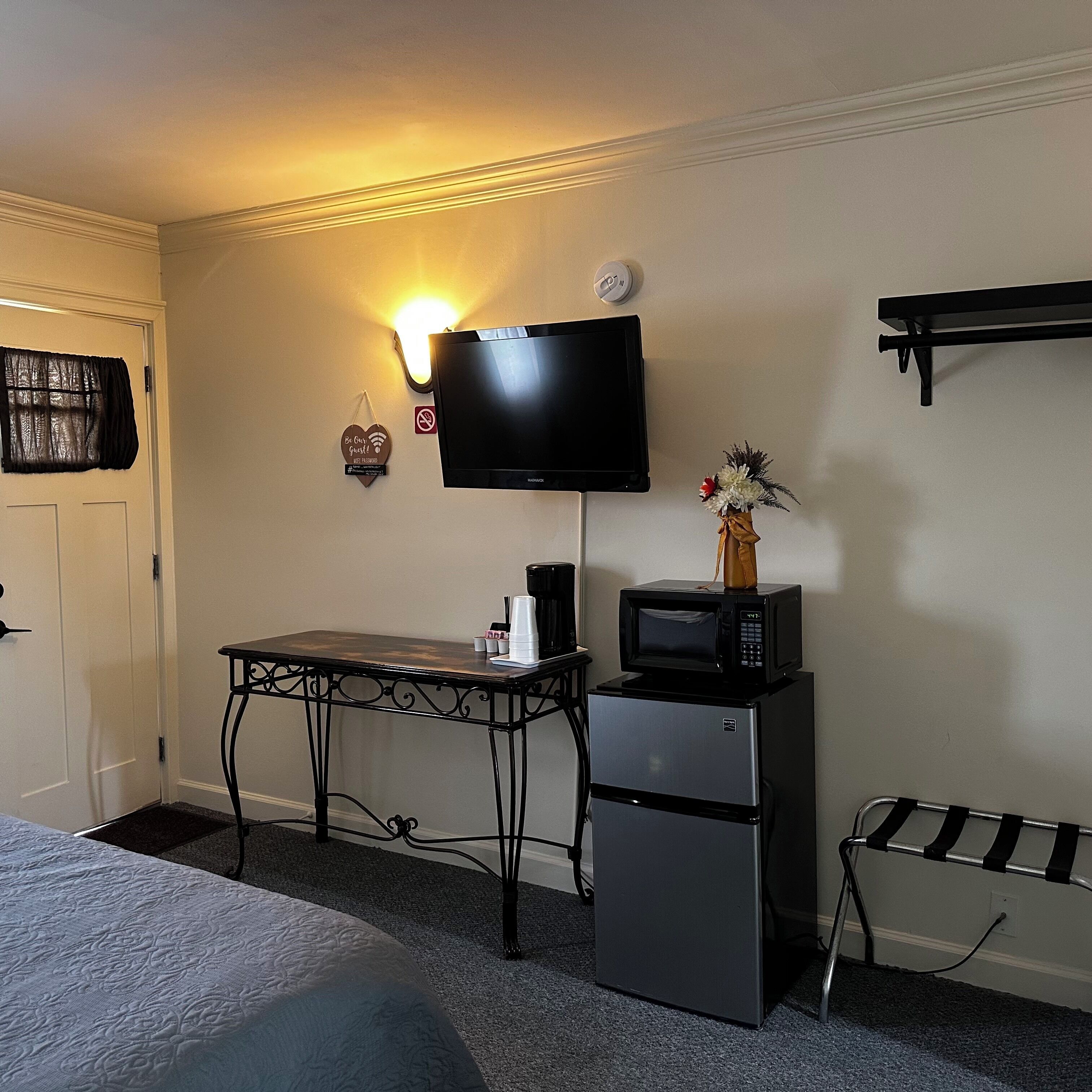 Ein kleines Hotelzimmer mit einem Bett, einem Schreibtisch mit Stuhl, einer Mikrowelle, einem Kühlschrank, einem an der Wand befestigten Fernseher und einem kleinen Tisch.