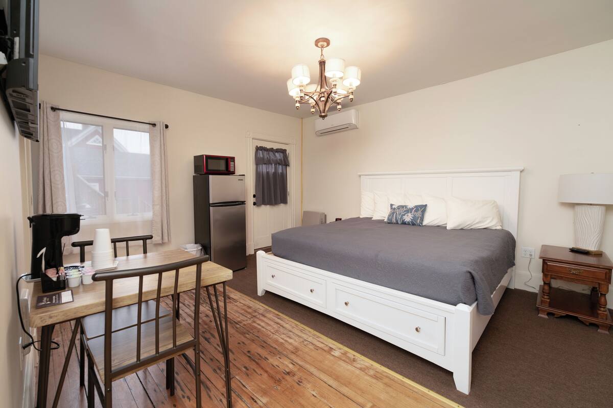 A bedroom with a bed, a desk with chairs, a microwave, and an air conditioner.
