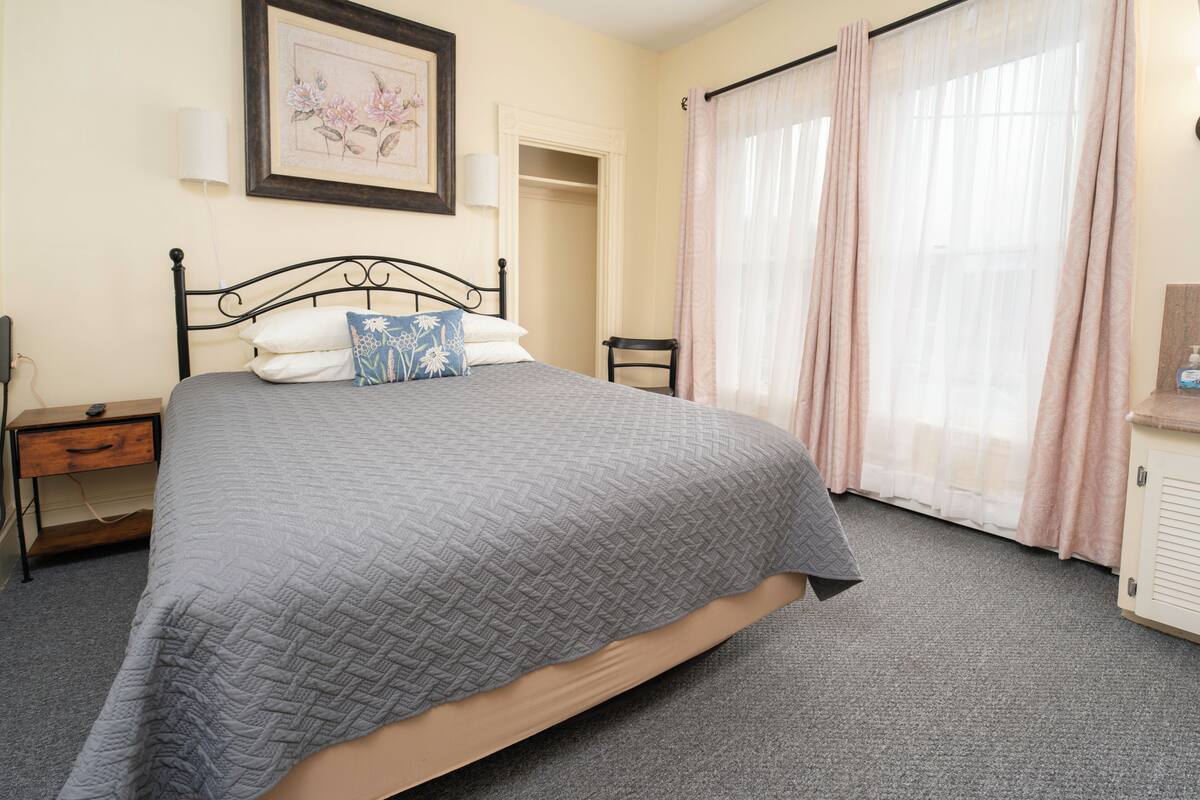 A bedroom with a bed, bedside table, chair, and a window with curtains.