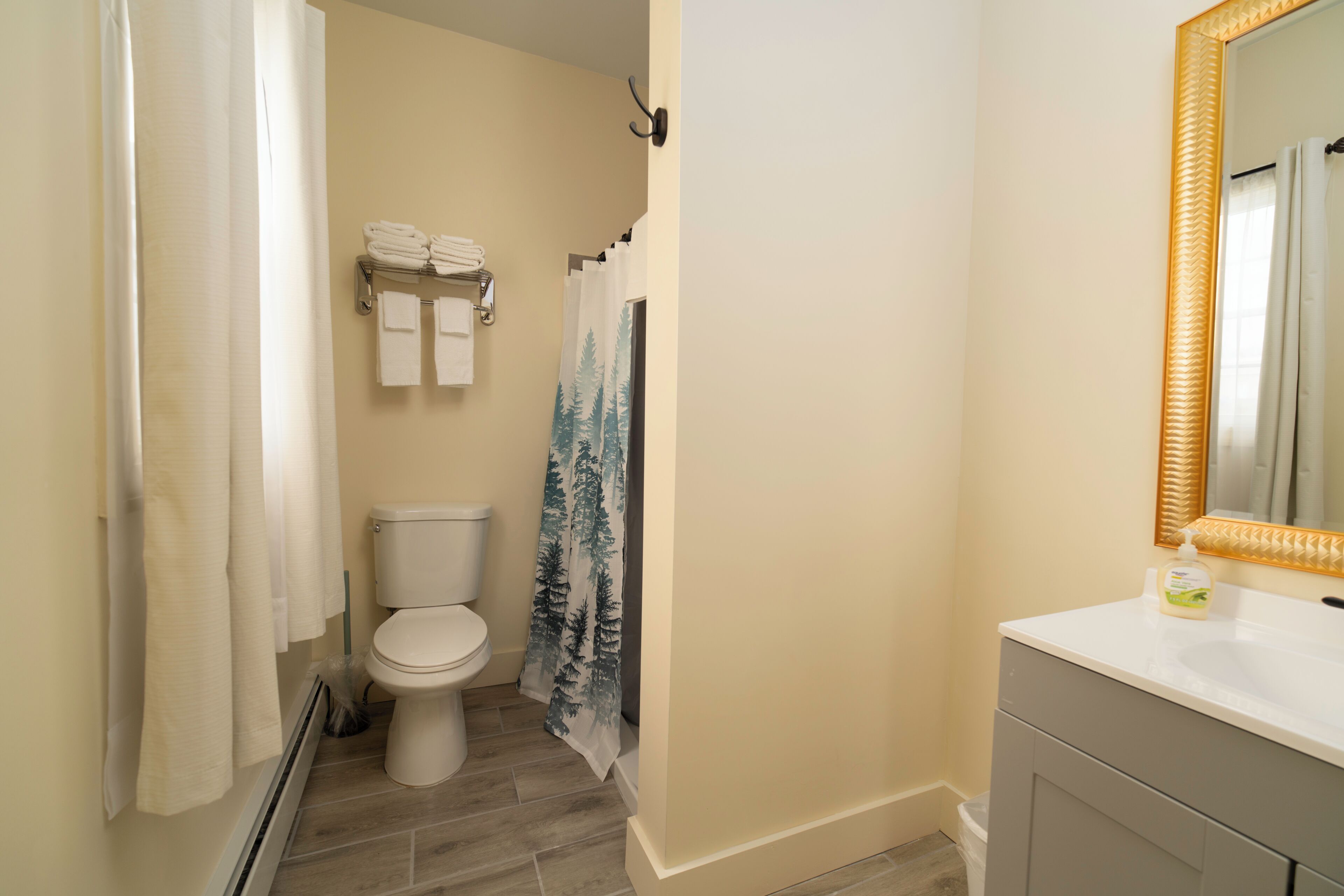 Une salle de bain comprenant des toilettes, un porte-serviettes avec des serviettes pliées, un rideau de douche orné d’un motif de forêt et un miroir avec un cadre doré.