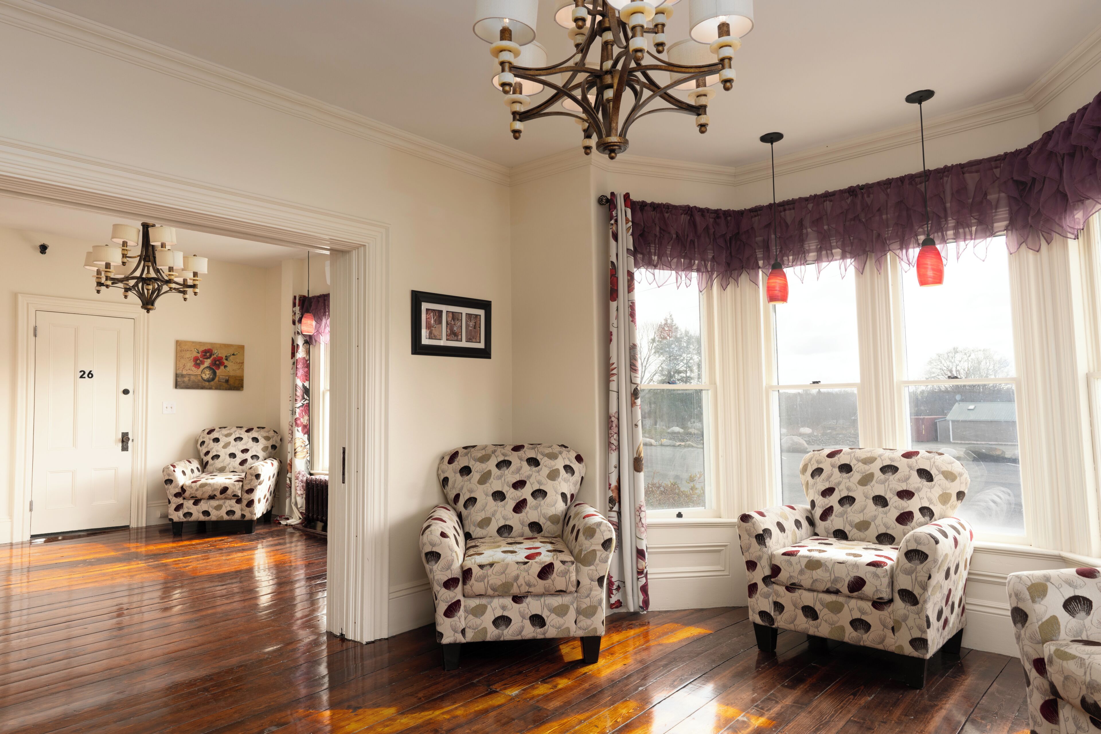 A room with a wooden floor, two armchairs with a polka dot pattern, a chandelier, and a window with curtains.