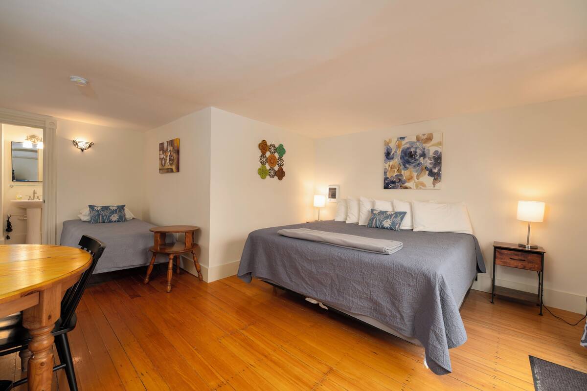 A hotel room with a bed, a desk, a chair, a small table, a lamp, and artwork on the walls.
