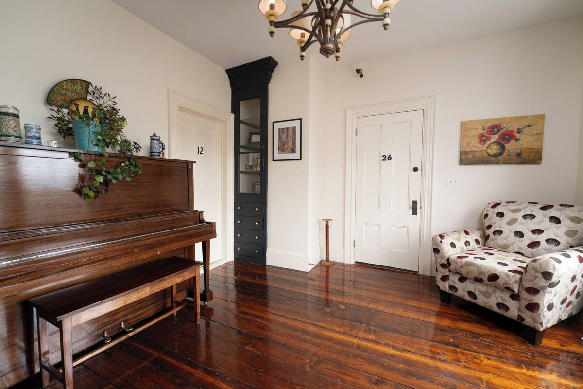 A room with a piano, a patterned armchair, and a door with numbers 12 and 26.
