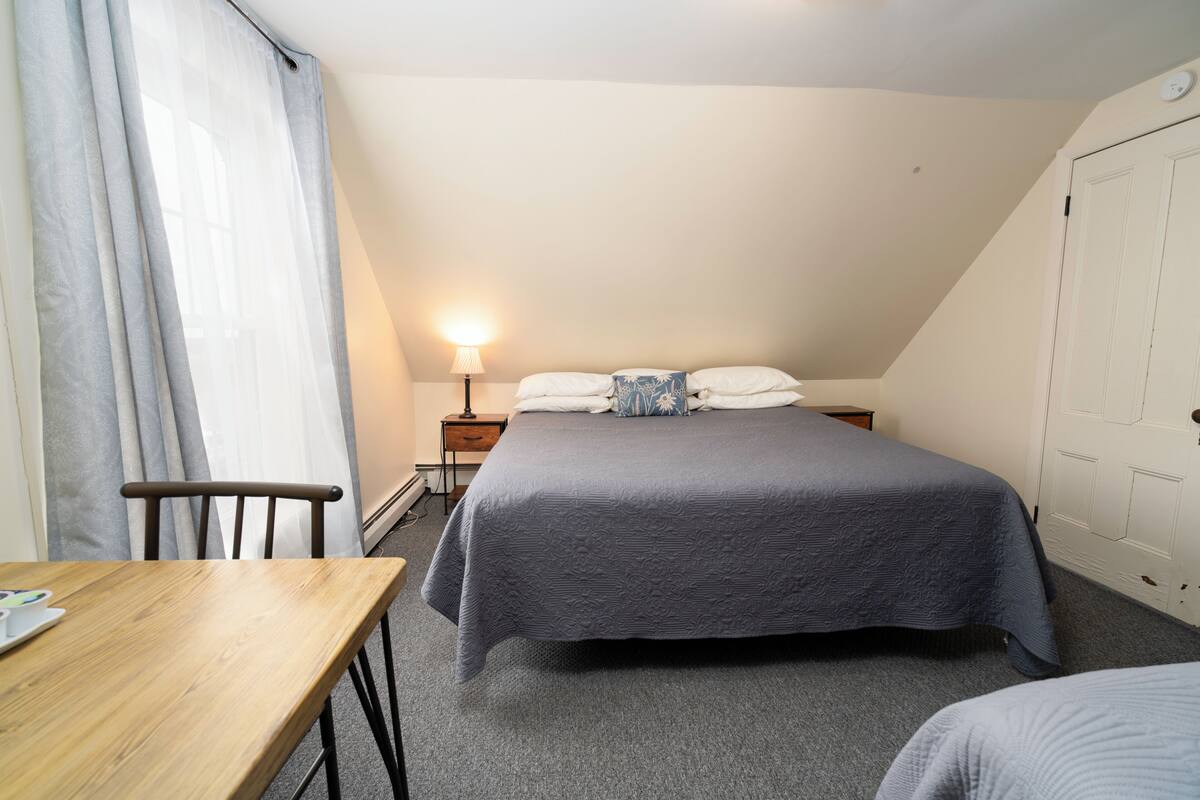 A bedroom with a bed, a bedside table, a lamp, a chair, and a window with curtains.