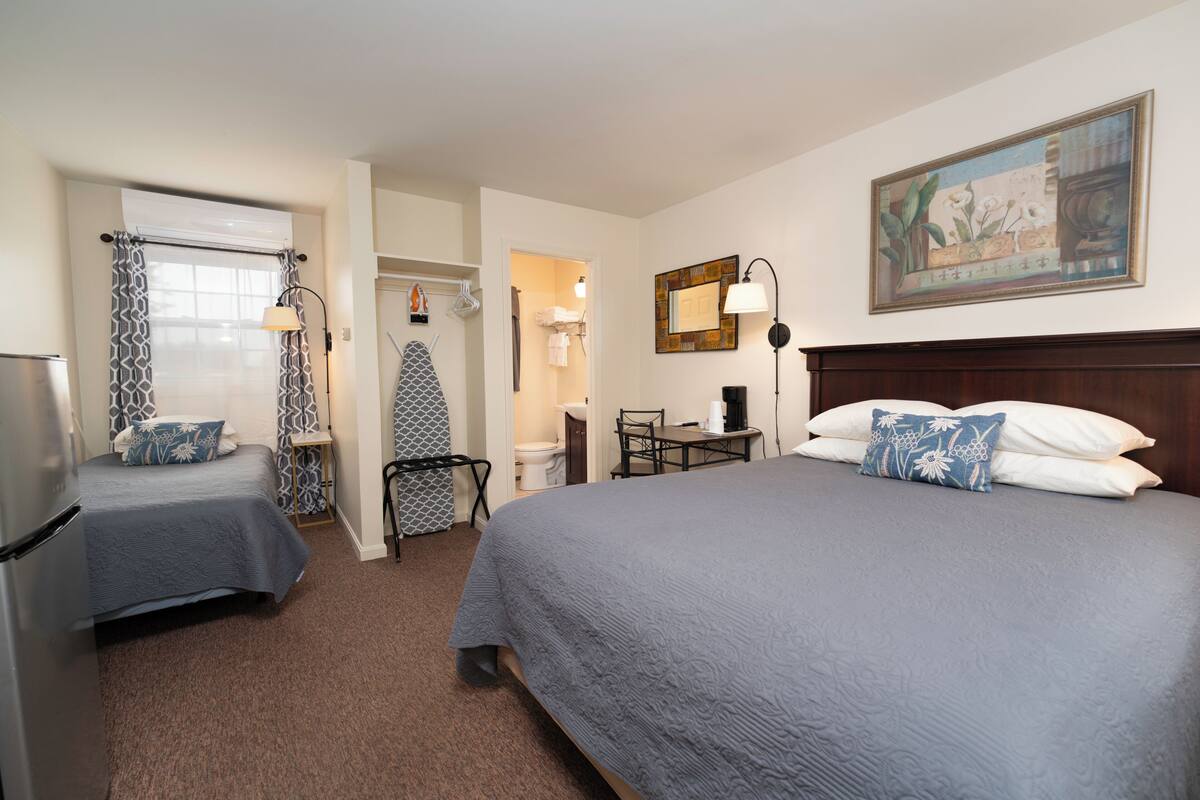 A hotel room with two beds, a desk, a chair, a refrigerator, a mirror, and a painting on the wall.