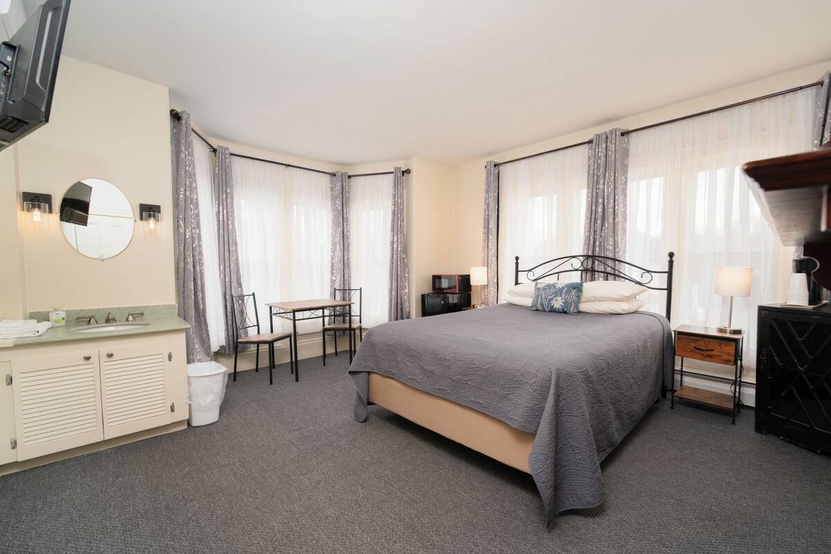 A hotel room with a bed, a desk, a chair, a TV, a lamp, a mirror, and a trash bin.