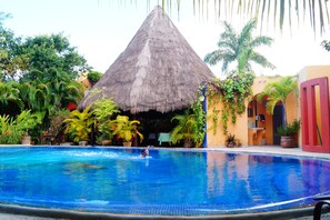 A swimming pool with a thatched-roof structure in the background, surrounded by lush tropical plants. - Casa Don Diego (Tulum)