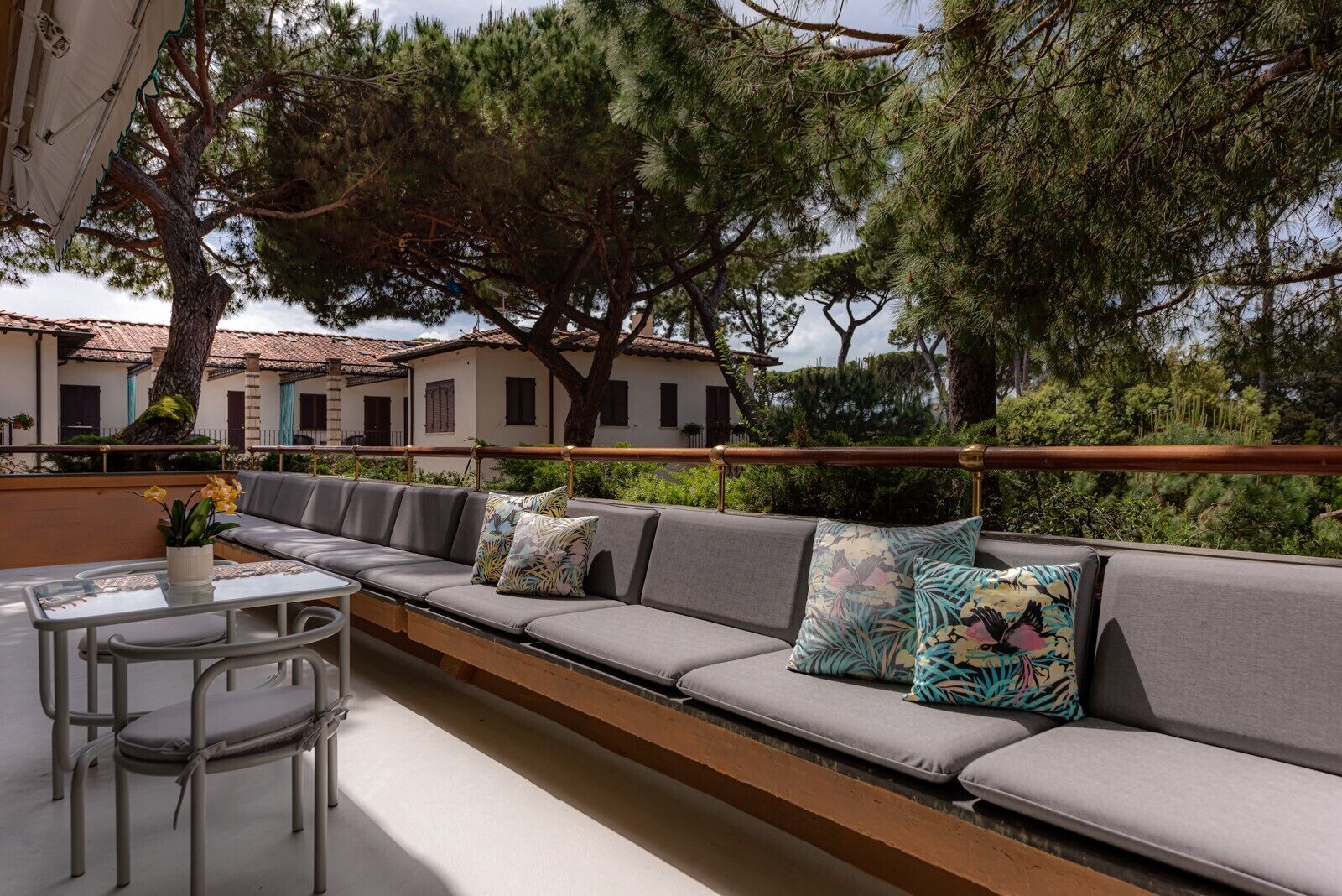 Un lungo divano grigio con cuscini floreali, un tavolino in vetro e sedie in metallo su un balcone con vista su edifici e alberi.