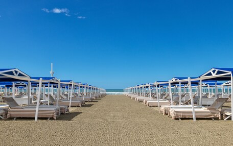 A beach with rows of sunbeds under blue umbrellas, a clear sky, and the ocean in the background.