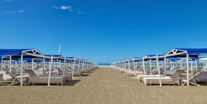A beach with rows of sunbeds under blue umbrellas, a clear sky, and the ocean in the background. - Augustus Hotel & Resort (Forte dei Marmi)