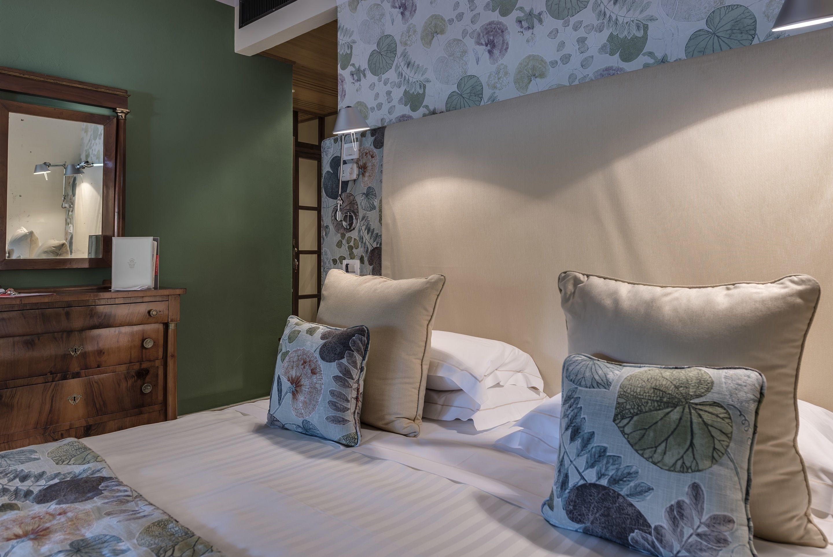 Une chambre à coucher avec un lit, deux oreillers, une commode, un miroir et un mur orné d’un motif floral.