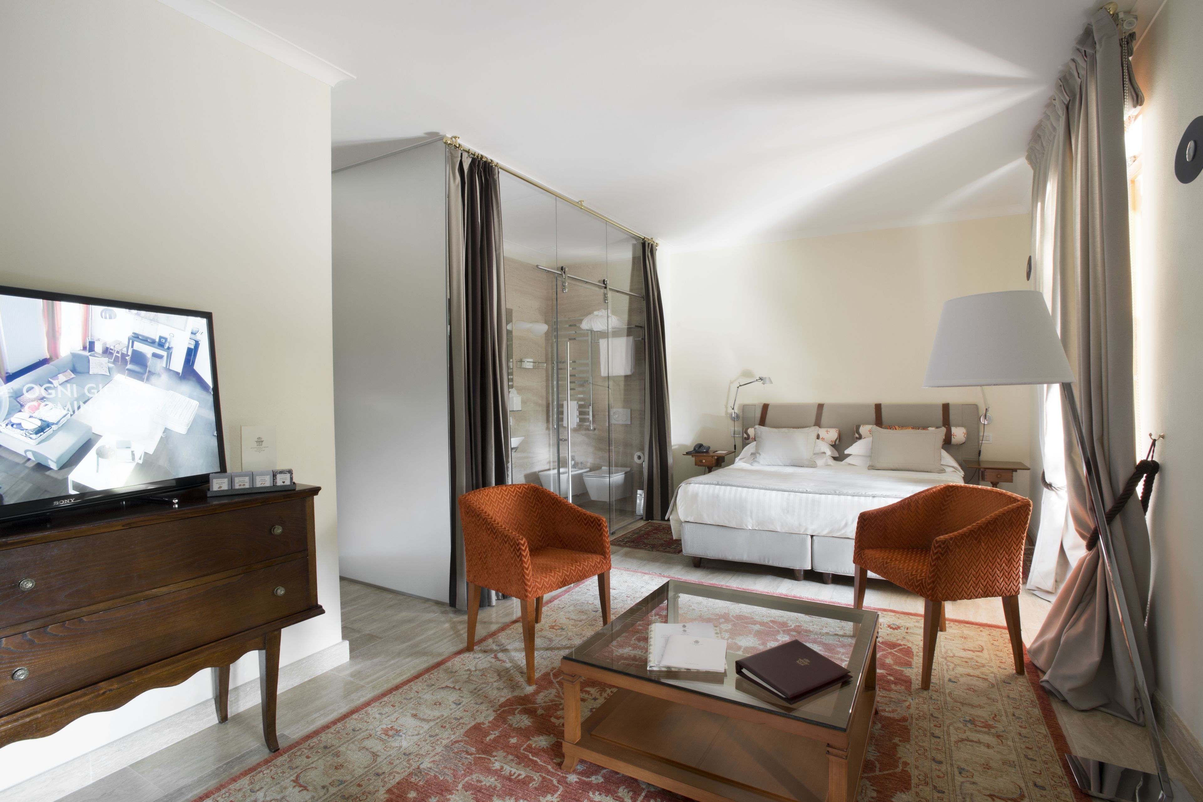 Una camera d'albergo con un letto, due poltrone, un tavolino, una TV e un box doccia in vetro.