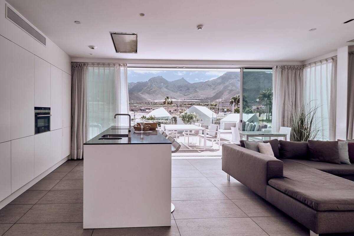 A modern kitchen with a central island, a dining area with a white table and chairs, and a large sofa. A view of mountains and a desert landscape is visible through the large windows.