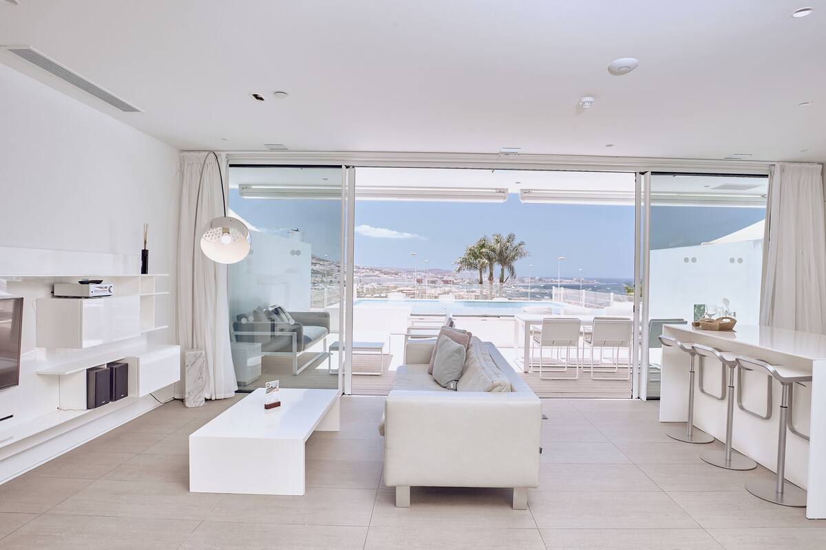 A modern living room with a sofa, coffee table, and a TV. There is a balcony with a view of the city and palm trees.