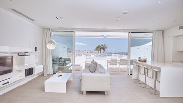 Un moderno salón con sofá, mesa de centro y televisor. Hay un balcón con vistas a la ciudad y palmeras.