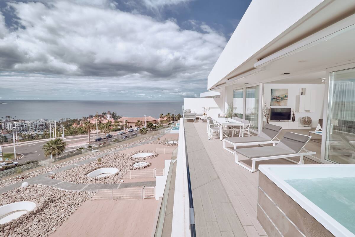 A modern balcony with a pool, lounge chairs, and a view of the sea and cityscape.