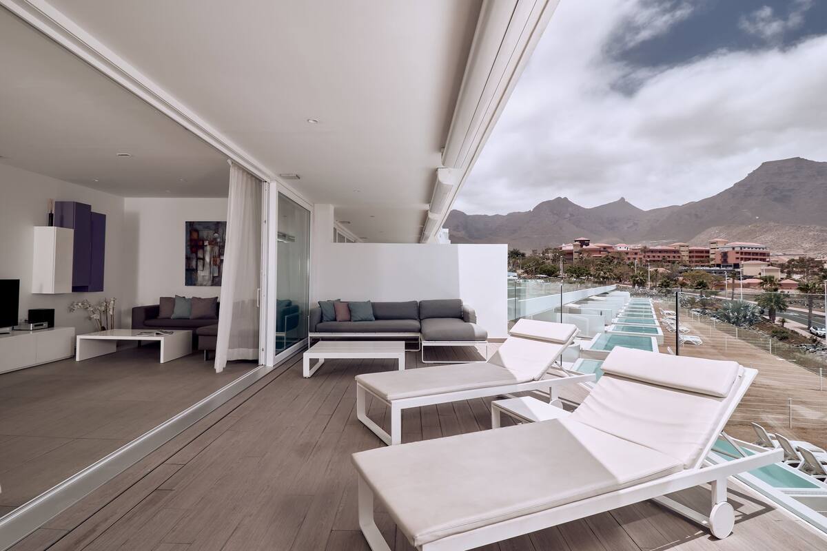 A modern living room with a sofa, coffee table, and lounge chairs. A large window offers a view of mountains and a pool area.